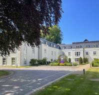 White multi-story building with black roof, surrounded by green lawn, trees, and a driveway under a clear blue sky.