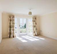 Empty room with cream-colored walls, beige carpet, floral curtains, and glass doors letting in sunlight from an outside garden.