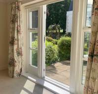 Open glass door with floral curtains leads to a sunlit patio and garden with green bushes and a metal garden arch outside.
