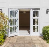 A pair of glass double doors open outward from a white building onto a stone patio, with green shrubs and a small tree on either side.