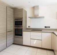 Modern kitchen with light wood cabinets, built-in double ovens, electric cooktop, stainless steel hood, and beige countertops in an L-shaped layout.