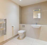 Modern bathroom with beige tiles, a wall-mounted toilet, a towel radiator, a wall mirror above a small sink, and a partial view of a bathtub.