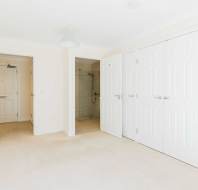 A bright, empty room with beige carpet, three white closet doors, and an open door leading to a tiled bathroom with a shower.