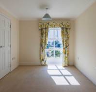 Empty room with beige walls and carpet, double closet doors on the left, and a window with floral curtains letting in sunlight.