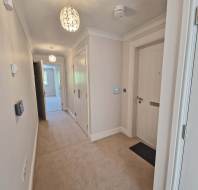 A well-lit hallway with beige carpet, white walls, ceiling lights, a closed front door, and doors leading to other rooms.