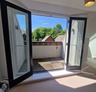 Open glass doors lead to a small balcony with a partial view of rooftops and trees in the background under a clear blue sky.