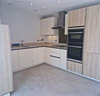 Modern kitchen with light wood and white cabinets, built-in oven and microwave, electric cooktop, sink, and tiled floor in an L-shaped layout.