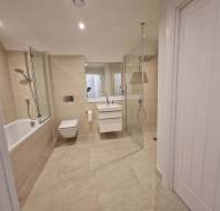 Modern bathroom with beige tiles, a wall-mounted toilet, bathtub with glass screen, walk-in shower, white vanity with sink, and large mirror.