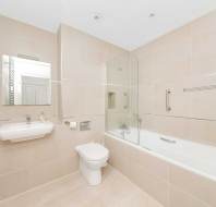 Modern bathroom with beige tiles, featuring a wall-mounted sink, toilet, bathtub with glass screen, mirror, and chrome towel rail.