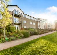 A modern, multi-story residential building with balconies and landscaped gardens, photographed on a sunny day with a clear blue sky.