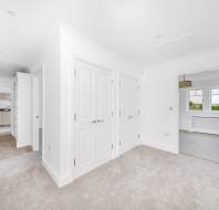 A bright hallway with white walls, beige carpet, multiple doors, and an open view into a kitchen and a room with a large window.