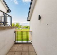 A small balcony with tiled flooring and metal railings overlooks a grassy area with trees and blue sky in the distance.
