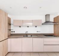 Modern kitchen with light wood cabinets, beige drawers, built-in ovens, electric cooktop, stainless steel extractor hood, and a sink under white upper cabinets.