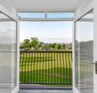 Open glass doors leading to a balcony with a black railing, overlooking a green lawn and trees under a partly cloudy sky.