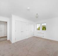 Bright, empty room with beige carpet, white walls and ceiling, built-in wardrobes, a window, and an open door leading to a hallway.