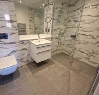 Modern bathroom with marble-patterned wall tiles, a wall-mounted toilet, floating sink vanity, large mirror, and walk-in glass shower enclosure.