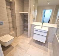 Modern bathroom with a wall-mounted toilet, glass shelves, heated towel rail, white sink vanity, large mirror, and a bathtub with a glass screen.
