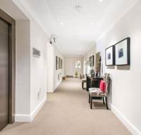 A modern hallway with beige walls, carpeted floor, an elevator on the left, and decorative chairs, art, and mirrors along the right wall.