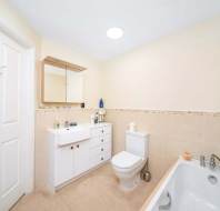 A clean, modern bathroom with a white vanity, mirror cabinet, toilet, and bathtub with a handheld showerhead against beige tiled walls.