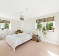 A bright bedroom with a double bed, striped bedding, two bedside tables, a chair with items on it, houseplants, and large windows with patterned blinds.