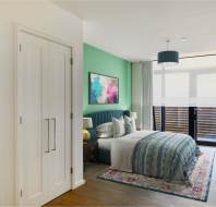 A modern bedroom with a green accent wall, a bed with blue and white bedding, bedside tables, a colorful rug, and glass doors leading to a balcony.