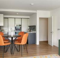 Modern kitchen and dining area with orange chairs around a round table, white cabinets, wooden floor, and a green sofa partially visible on the right.