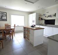 Bright kitchen with white cabinets, a central island, wooden dining table with chairs, tiled floor, and a large window letting in natural light.