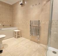 A bathroom with a bathtub, shower, toilet, sink, towel warmer, small stool, and beige tiled walls and floor.