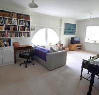 A bright living room with a bookshelf, desk, sofa, armchairs, TV, piano, and large window with an arched section.