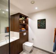 Modern bathroom with a wall-mounted toilet, wooden shelving with towels and decor, a sink with mirror, and a framed picture on a white wall.