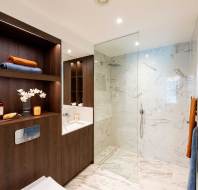 Modern bathroom with wood cabinetry, a small sink, decorative items, and a walk-in glass shower with marble walls and floor. Towels are neatly displayed on shelves and a rack.