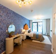 Modern bedroom with a blue patterned accent wall, a bed, a vanity table with a mirror, and large windows letting in natural light.