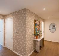 A hallway with patterned wallpaper, a white door, a marble console table with a vase, a colorful portrait, and an oval mirror on a beige wall.