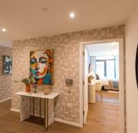 A hallway with patterned wallpaper, a marble console table, and a colorful abstract portrait; a bedroom is visible through an open door.