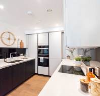 Modern kitchen with white countertops, black cabinets, built-in appliances, decorative jars and plants, and a wooden clock on the wall.
