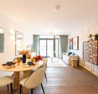 A modern open-plan living and dining area with light wood flooring, a round dining table, upholstered chairs, a patterned cabinet, and large windows letting in natural light.