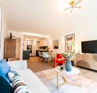 Bright, modern living room with a sofa, coffee table, TV, dining area, and open kitchen. Decor includes framed art, plants, and contemporary furniture.