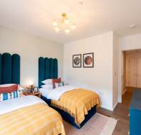 A modern bedroom with two single beds, upholstered blue headboards, yellow blankets, framed artwork on the wall, and a pendant light fixture.
