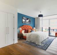 Modern bedroom with an orange upholstered headboard, blue accent wall, white bedding, patterned rug, armchair, two nightstands, and large window with sheer curtains.