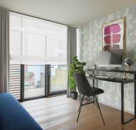 A modern home office with a glass desk, computer, chair, indoor plant, and large window with sheer curtains. A blue sofa sits to the left with striped pillows.