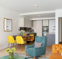 Modern open-plan living and dining area with teal and orange seating, yellow dining chairs, a kitchen in the background, and a wall-mounted mirror.