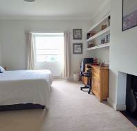 A bright bedroom with a double bed, wooden desk, office chair, shelves, fireplace, chest of drawers, and a window with light curtains.