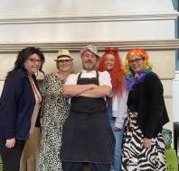 Five adults stand indoors in front of a fireplace, wearing various costumes and colorful wigs, posing and smiling for the camera.