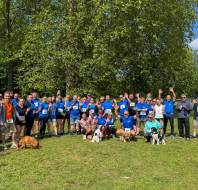 A large group of people, some with dogs, stand on grass in front of trees, wearing athletic clothes and race bibs, smiling and waving at the camera.
