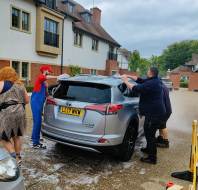 Three people, including one in a red wig and another in blue overalls and red cap, wash a silver SUV parked outside a residential building.