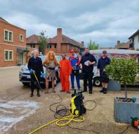 Six people stand outside near brick buildings, some in costumes including Mario and Chewbacca, with cleaning equipment and a foam-covered pressure washer in the foreground.