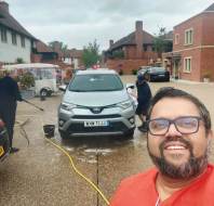 A man in glasses and a red shirt takes a selfie in front of people washing a white Toyota SUV in a residential area.