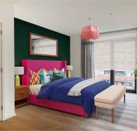 A modern bedroom with a pink bed, blue bedding, wooden nightstands, a large window with gray curtains, a potted plant, and a green accent wall with a mirror.