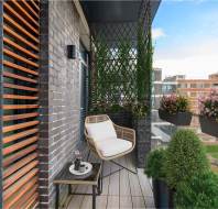A modern balcony with a chair, small table, potted plants, and a privacy screen, overlooking a cityscape with buildings and greenery.