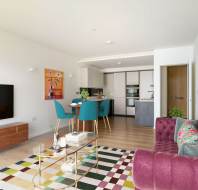 Modern open-plan living and kitchen area with teal dining chairs, a purple sofa, wall art, a large TV, and a colorful rug on light wood flooring.